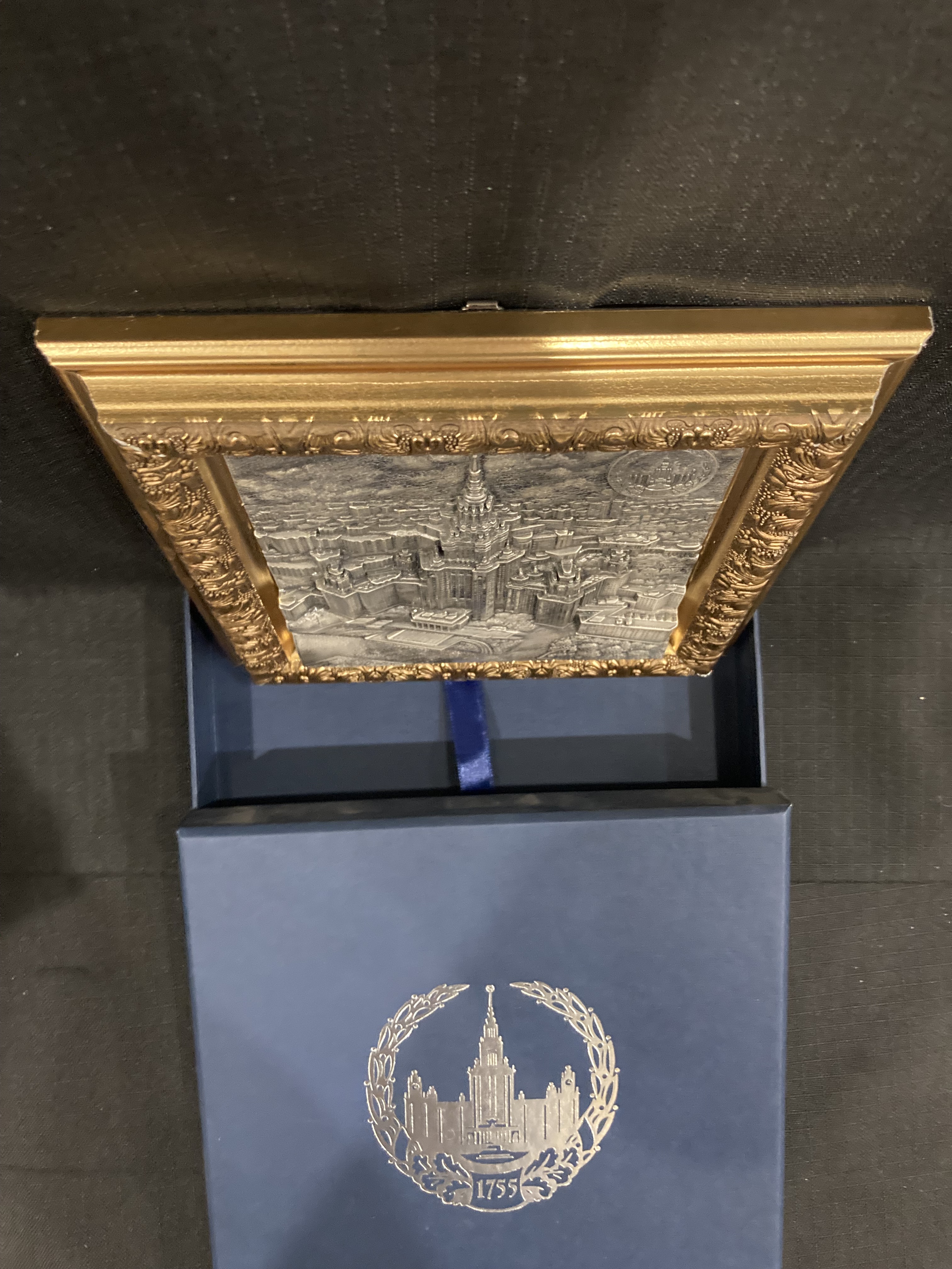 Продажа настенного рельефного панно размером 12,5x12,5 см с изображением здания Московского государственного университета имени М.В. Ломоносова (далее - МГУ) на Воробьевых горах и эмблемы МГУ с покрытием из серебра 960 пробы в багетной рамке золотистого цвета в подарочном футляре синего цвета с серебристой эмблемой МГУ в центре.