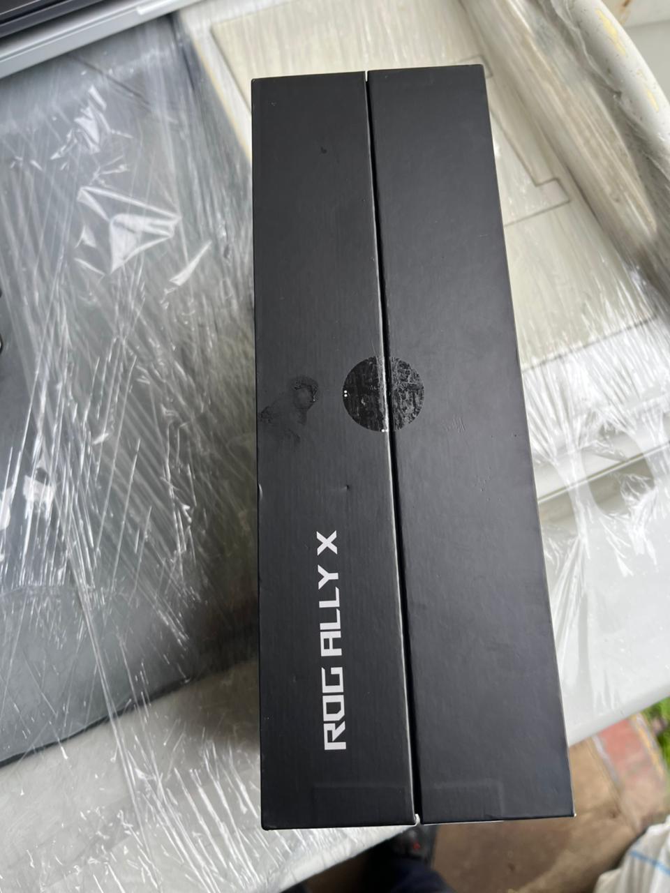 Картонная коробка черного цвета с рисунками и надписью ROG ALLY X, внутри коробки находится игровая консоль черного цвета