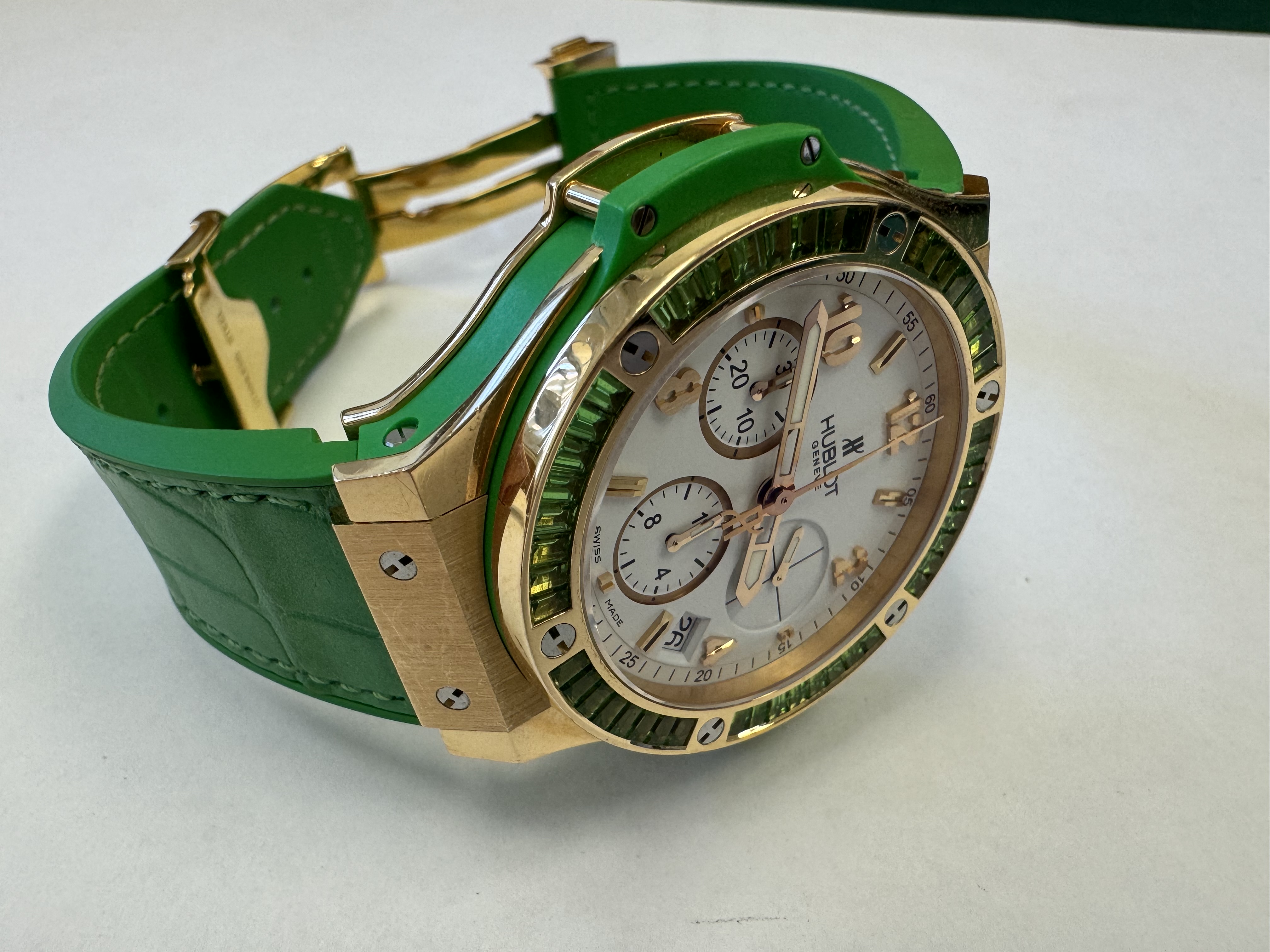 Наручные часы «Hublot Big Bang TuttiFrutti Apple ref. 341.PG.2010.LR.1922» в круглом корпусе из металла золотистого цвета с вставками зеленого цвета и камнями зеленого цвета по кругу. Часы имеют ремешок из материала зеленого цвета с замком из металла золотистого цвета. На оборотной стороне часов имеется штампованная надпись «HUBLOT», «BIG BANG», «341», «896995», «Gold 750», б/у.